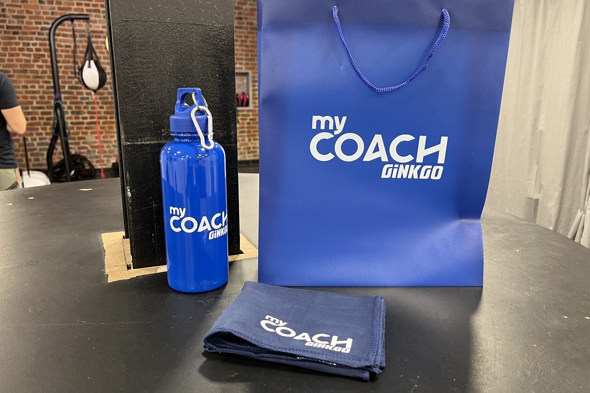 Une gourde, un sac et une serviette My Coach posés sur un banc.