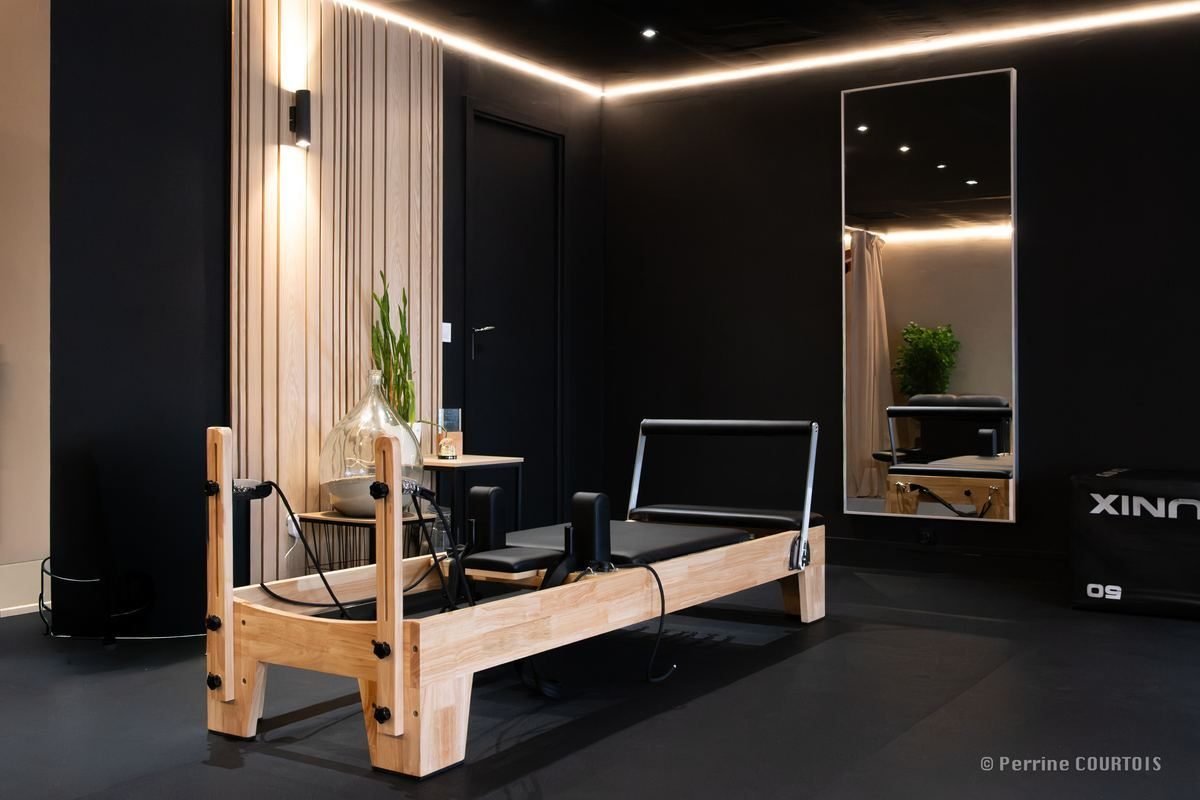 Un studio My Coach : reformer de Pilates en bois clair, miroir pleine hauteur et éclairage indirect sur murs sombres.
