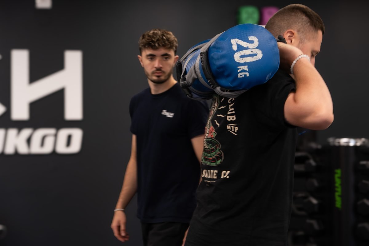 Un adhérent porte un sac lesté sur l'épaule sous le regard de son coach, au studio de Levallois-Perret.