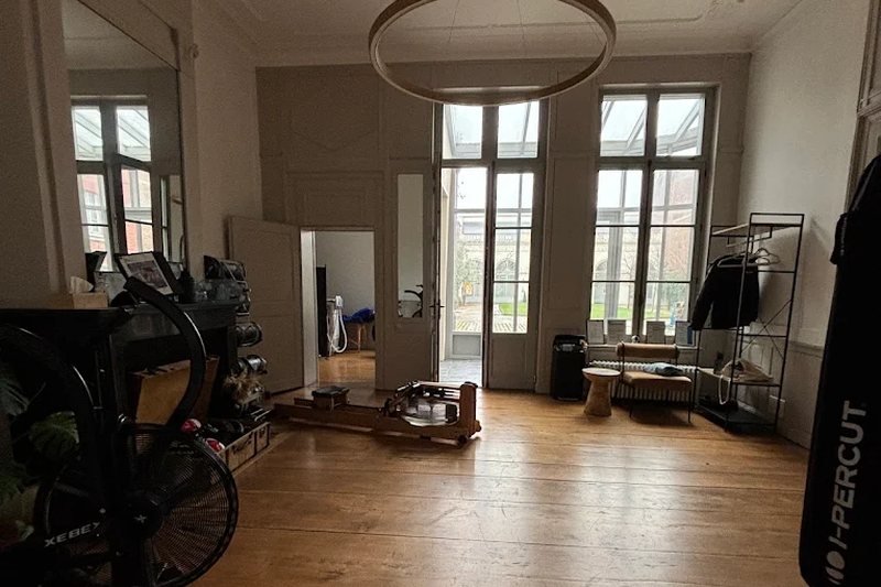 Vue d'ensemble d'une salle du studio de Lille : parquet, grandes fenêtres, vélo à air, rameur et sac de frappe.